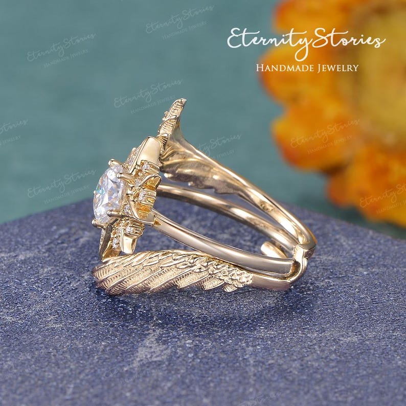 Puede incluir: Un anillo de compromiso dorado con una gran gema transparente en el centro. El anillo presenta detalles intrincados, incluido un dise&ntilde;o en forma de pluma en la banda y alrededor de la piedra central. El texto "Eternity Stories Handmade Jewelry" es visible.