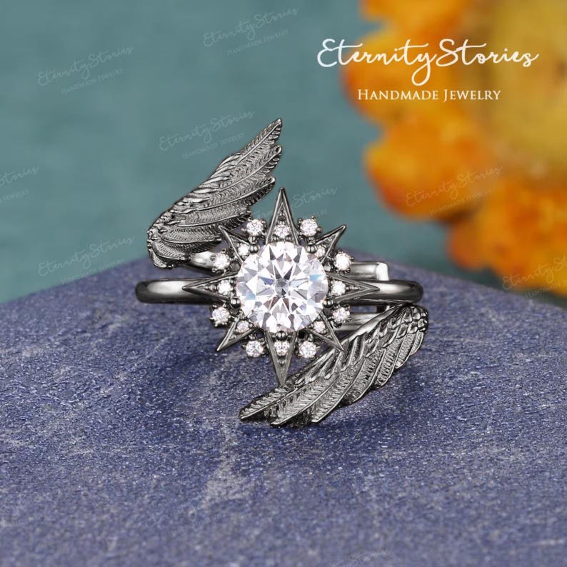 Puede incluir: Anillo hecho a mano con una piedra central grande y transparente rodeada de piedras m&aacute;s peque&ntilde;as en forma de estrella. El anillo presenta detalles de alas. El texto "Eternity Stories Handmade Jewelry" es visible.