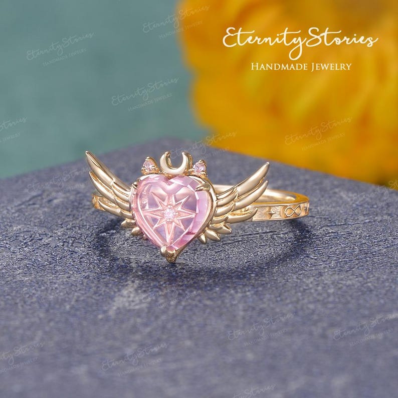 Pu&ograve; includere: Un anello color oro con una pietra preziosa rosa a forma di cuore con un disegno a stella. L'anello presenta accenti alati su entrambi i lati del cuore, una luna crescente e piccole gemme. La fascia ha un simbolo di infinito. Sullo sfondo c'&egrave; la scritta "Eternity Stories Handmade Jewelry".