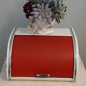 May include: A retro red and white bread bin with a curved top and a silver handle. The bread bin has a white top and a red front. A white ceramic vase with faux succulents and flowers sits on top of the bread bin.