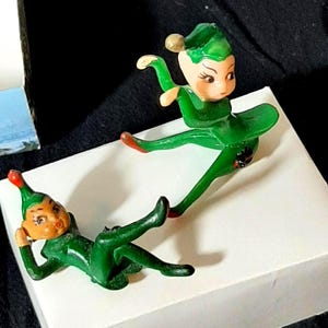 Puede incluir: Dos figuras vintage de duendes navideños. Los duendes visten trajes verdes con sombreros a juego y detalles rojos en los zapatos. Un duende está reclinado y el otro en una pose juguetona. Las figuras están sobre una superficie blanca.