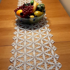 May include: A white, open-weave crochet table runner with a scalloped edge, displayed on a wooden table. A glass bowl filled with grapes, oranges, and other fruits sits atop the runner, adding a touch of colour.