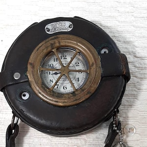 May include: A vintage Detex Newman watch in a round, dark brown leather case. The watch face has a brass bezel and a white dial with black numerals. Two keys hang from a chain attached to the case.