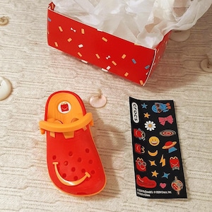 May include: A red box with a white interior and confetti patterns, filled with white tissue paper. Beside it is a red and orange clog-style shoe with a smiling face, and a sheet of Crocs stickers.