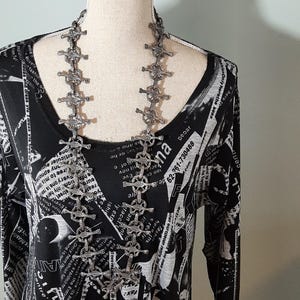 K&ouml;nnte beinhalten: Ein schwarz-wei&szlig; gemustertes Kleid mit einer silbernen Kettenkette mit Kreuzdesign. Die Kette hat mehrere kreuzf&ouml;rmige Glieder.