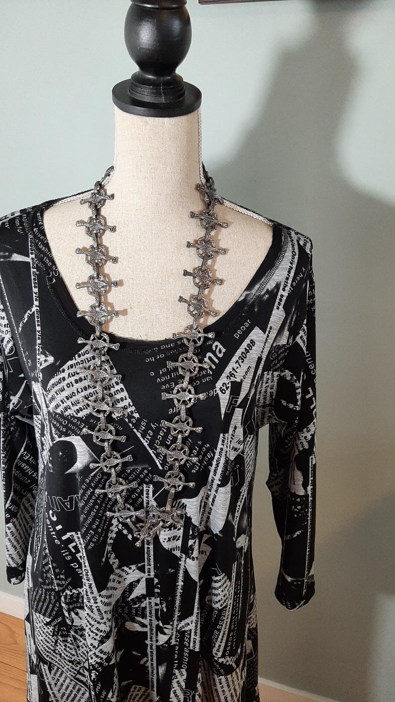 K&ouml;nnte beinhalten: Ein schwarz-wei&szlig; gemustertes Kleid mit einer silbernen Kettenkette mit Kreuzdesign. Die Kette hat mehrere kreuzf&ouml;rmige Glieder.
