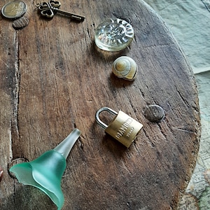 Puede incluir: Una superficie redonda de madera con una variedad de objetos sobre ella, incluyendo una llave, un pisapapeles de vidrio, una concha, un candado y un embudo de vidrio verde. El candado es dorado y tiene la palabra "DIAMOND" y el número "76002" en él.