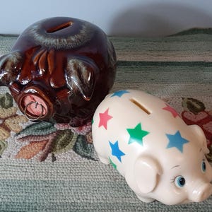 Pode incluir: Dois cofrinhos de cerâmica. Um é marrom com acabamento envidraçado e o outro é branco com estrelas coloridas.