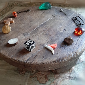 Puede incluir: Una superficie circular de madera con varios objetos dispuestos alrededor, incluyendo una calavera, una cruz, una llave, una espada, un trozo de vidrio verde, una cuenta de vidrio roja, un anillo de metal, un pequeño disco de madera, un dado negro con una calavera en él, un diente blanco con un borla roja y una roca marrón.