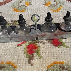 May include: Four small, clear glass bottles with black stoppers, set in a weathered, rectangular metal holder. A small metal handle is between two of the bottles. The bottles are on a patterned, off-white textile.