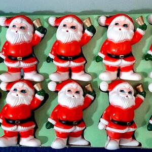 Puede incluir: Una colección de diez figuras vintage de Papá Noel. Cada figura viste un traje rojo con cinturón negro, barba blanca y una campana en la mano. Las figuras están dispuestas sobre un fondo verde, ideales para la decoración navideña.