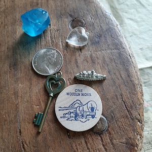Könnte beinhalten: Ein Holztoken mit blauem und weißem Text, der "ONE WOODEN NICKEL" lautet, und einer blauen und weißen Illustration eines Planwagens. Der Token liegt auf einer Holzoberfläche mit einem silbernen Schlüssel, einer Silbermünze, einem blauen Glasstück und einem klaren Glasherz.