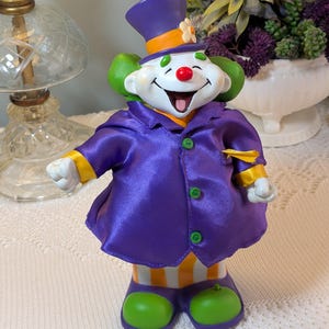 Peut inclure: Une figurine de clown portant un haut-de-forme violet avec une bande orange et une fleur blanche, une veste violette brillante à boutons verts et des poignets jaunes. Le clown a des cheveux verts, un nez rouge et un pantalon rayé orange et blanc.
