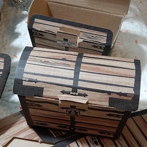 May include: A collection of cardboard boxes designed to resemble treasure chests. The boxes feature a printed wood grain pattern in shades of brown with black accents representing metal straps, hinges, and a central latch mechanism.