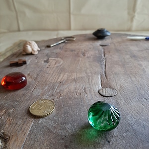 Puede incluir: Una superficie de madera con una variedad de objetos, incluyendo una pequeña figurita de cerámica, una cuenta de vidrio roja, una moneda de oro y una cuenta de vidrio verde.