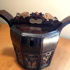 Könnte beinhalten: Dunkelbraune, antike Holzkiste mit Klappdeckel und geschnitzten Paneelen. Die Kiste hat eine einzigartige Form mit geschwungenen Griffen und goldfarbenen Drachenakzenten. Die Kiste ist achteckig.