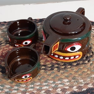 Puede incluir: Una tetera de cerámica marrón y dos tazas con un diseño de cara tribal. La tetera tiene una cara verde, amarilla, roja y blanca con una boca y dientes blancos. Las tazas tienen un diseño similar con caras verdes, amarillas, rojas y blancas.