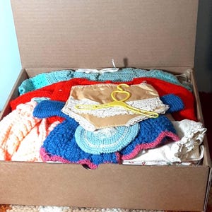 Könnte beinhalten: Ein Karton voller Vintage-Kleidung. Der Karton enthält eine Auswahl an farbenfrohen Strick- und Häkelkleidungsstücken, darunter Pullover, ein blau-rosa gehäkeltes Teil und Unterwäsche in Beige mit Spitzenbesatz. Ein gelber Kleiderbügel liegt obenauf.