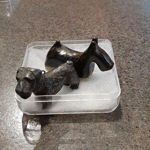 Puede incluir: Dos pequeñas figuras de perro. Una es una escultura detallada de metal oscuro de un terrier. La otra es un perro de cerámica suave, marrón oscuro, con cuello curvo. Ambas se exhiben en un recipiente de plástico transparente.