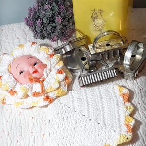 May include: A selection of vintage kitchenware, featuring a yellow canister, metal cookie cutters, and a crocheted doll head cover. The doll head cover is white with yellow and orange detailing. A white crocheted potholder with yellow and orange trim is also visible.