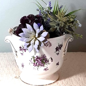 Puede incluir: Un jarrón de cerámica blanco con base de pedestal y dos asas, adornado con patrones florales morados y ribetes dorados. El jarrón está lleno de flores artificiales en tonos morados, verdes y blancos, creando un arreglo decorativo.