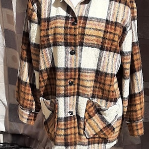 Puede incluir: Chaqueta vintage a cuadros en tonos marrón, crema y naranja, exhibida en un maniquí. La chaqueta presenta un cuello, cierre de botones y dos bolsillos delanteros. La tela tiene una apariencia texturizada.