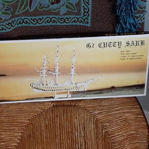 Peut inclure: Une boîte pour un kit de modèle de navire G2 Cutty Sark. La boîte montre un modèle de navire blanc avec une base en bois brun. Le texte sur la boîte indique "G2 Cutty Sark" et comprend des détails sur le navire, tels que son année de construction, son type, sa longueur et sa largeur.