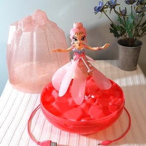 Könnte beinhalten: Eine rosa Puppe mit rosa Kleid und roter Basis. Die Puppe hat rosa Haare und steht auf einer roten, runden Basis. Ein rosa USB-Kabel ist mit der Basis verbunden. Ein rosa, geometrisch geformtes Objekt befindet sich hinter der Puppe.