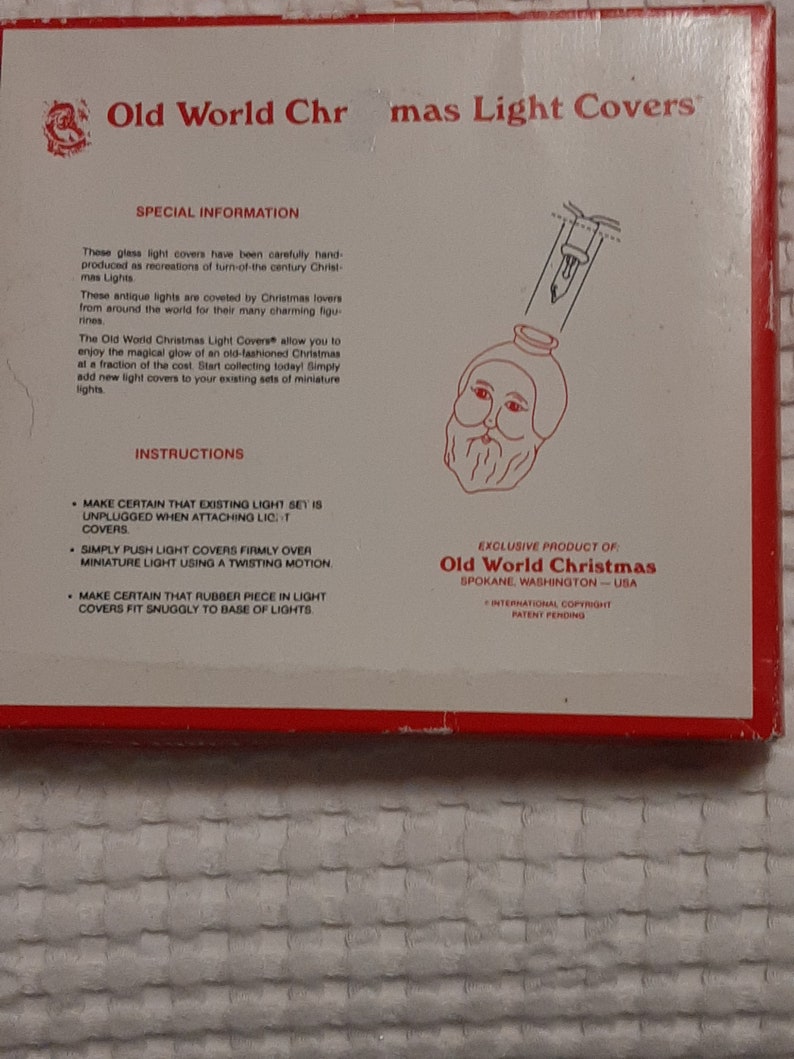 May include: A white box with red trim and the text "Old World Christmas Light Covers" printed on the front. The box features a black and white illustration of a Santa Claus light cover. The text "Exclusive Product of: Old World Christmas Spokane, Washington - USA" is printed on the bottom of the box.