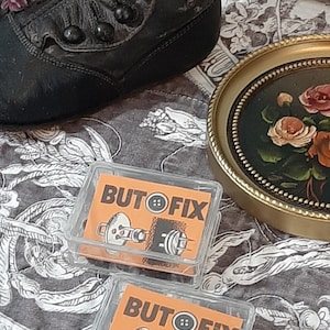 Könnte beinhalten: Ein antiker schwarzer Lederstiefel mit weißen Blumen im Inneren, neben zwei transparenten Plastikboxen mit orangefarbenen Etiketten mit der Aufschrift "BUTOFIX". Ein kleines gerahmtes Gemälde und ein Blumengemälde in einem goldenen Rahmen sind ebenfalls zu sehen.