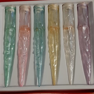 May include: Six iridescent glass bottle stoppers in a red and white box. The stoppers are in shades of green, pink, blue, yellow, white, and purple.