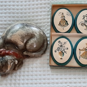 Op de afbeelding: Een set van vier vintage decoratieve keramische tegels met een teal rand. Elke tegel heeft een witte achtergrond met een zwart silhouetontwerp. De ontwerpen omvatten een vrouw in een jurk die een mand vasthoudt, een vrouw die aan een bureau zit, een vaas met bloemen en een vrouw in een jurk die een mand vasthoudt.