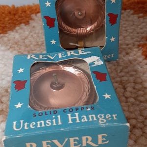 Może przedstawiać: Dwa zabytkowe wieszaki na przybory kuchenne z miedzi w oryginalnych niebieskich pudełkach. Pudełka zawierają nazwę "REVERE" i słowa "Solid Copper Utensil Hanger". Każdy wieszak jest widoczny przez przezroczyste okienko.