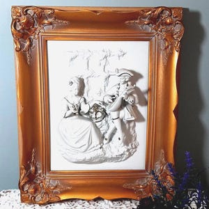 May include: A gold-framed relief sculpture of a couple in period dress, with a man playing a stringed instrument. The relief is white, set against a white background. The frame has an ornate design.