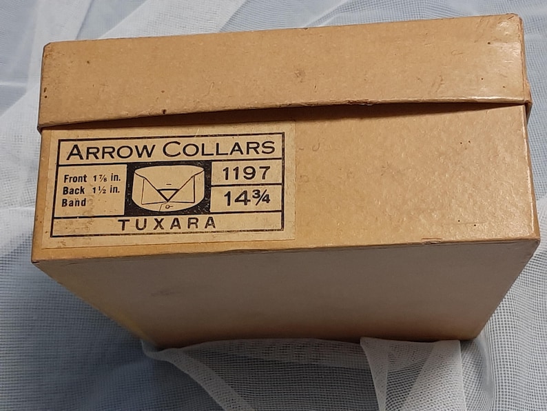 May include: A brown cardboard box with a white label that reads "ARROW COLLARS" and includes size information: "Front 1 1/8 in. Back 1 1/2 in. Band 14 3/4" and "TUXARA".