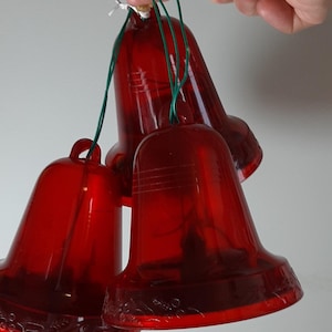 May include: Three transparent red bell ornaments suspended by green string. The bells have a traditional shape with a flared base and a loop for hanging. The bells are made of a shiny, translucent material, perfect for festive decorations.