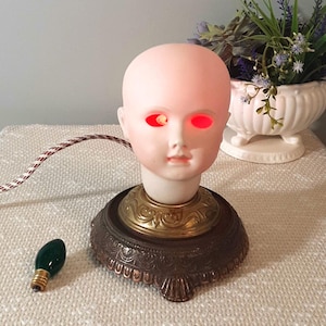 May include: A vintage doll head lamp with glowing red eyes. The doll head is mounted on a decorative bronze base with a brass accent. A green light bulb sits next to the lamp, and a patterned cord extends from the lamp.