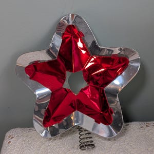 May include: A star-shaped tree topper featuring a shiny silver metal frame and a vibrant red metallic centre insert. The star has a five-pointed design with a coiled metal spring base attached to the bottom for mounting on a tree branch.