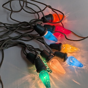May include: A string of vintage-style Christmas lights featuring large, textured, multi-coloured bulbs in orange, pink, blue, yellow, and green. Each bulb is set in a dark, matte-finished socket and connected by a thick, braided brown fabric cord.