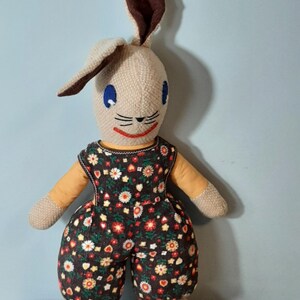 Könnte beinhalten: Eine braune und beige gestrickte Hasenpuppe, die eine braune Latzhose mit Blumenmuster trägt. Die Puppe hat blaue Augen und ein rotes Lächeln.