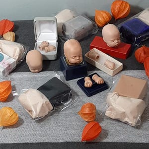 Puede incluir: Surtido de partes de muñecas en varias etapas de finalización, incluyendo cabezas, brazos y piernas. Las partes se muestran en pequeñas cajas y bolsas sobre una superficie con estampado gris y blanco. Algunas de las partes están hechas de un material marrón claro, mientras que otras están hechas de un material blanco. También hay varias pequeñas linternas de papel naranja esparcidas alrededor de las partes.