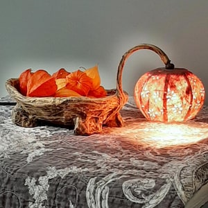 Könnte beinhalten: Eine Holzschale gefüllt mit orangefarbenen Papierlaternen steht auf einer gemusterten Tischdecke neben einer beleuchteten Lampe aus einer getrockneten Kalebasse.