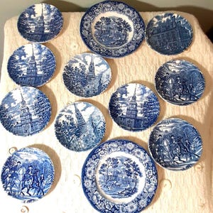Può includere: Una collezione di piatti decorativi blu e bianchi. I piatti presentano varie scene storiche, tra cui edifici e figure a cavallo. I piatti sono disposti su una superficie color crema, mettendo in risalto i loro intricati disegni e motivi.
