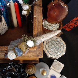 May include: A wooden box with a white, blue, and yellow crystal wand with a deer antler handle. The wand is decorated with moss, seashells, and crystals.
