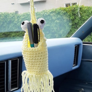 May include: A yellow crocheted octopus car air freshener with two black eyes and two pens sticking out of its head. The octopus is hanging from the rear view mirror of a car.