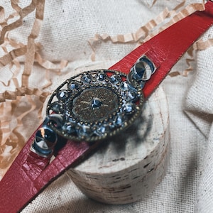May include: A red leather bracelet featuring a large, circular silver charm. The charm is adorned with rhinestones and a floral design.