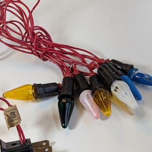May include: A string of vintage Christmas lights featuring a red electrical cord and black sockets. The bulbs are a mix of colours including yellow, green, pink, white, and blue, with some showing paint wear. A tag reads Approved SP Standard.