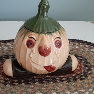 May include: A ceramic pumpkin cookie jar with a green stem and a painted face. The pumpkin has a wide smile, red eyes, a red nose, and red tears. The pumpkin is sitting on two brown legs.