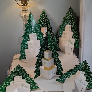 May include: A collection of green cardboard Christmas tree decorations featuring white star patterns and attached white gift box cutouts. Each tree has a flat base and includes printed text labels such as A and B on the bottom sections.