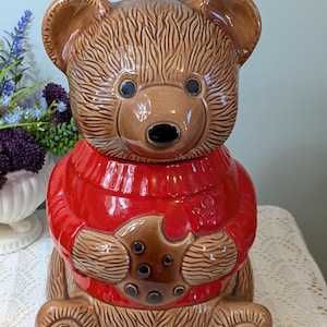 Peut inclure: Un pot à biscuits en céramique marron en forme d'ours, vêtu d'un pull rouge, tenant un biscuit avec une bouchée. L'ours a des yeux noirs et un nez noir. Le pot est sur une nappe en dentelle blanche.
